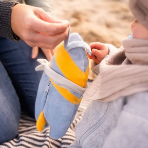 A child gently holds Nalu, a soft surfboard-shaped plushie in light blue with bright yellow accents and a white band detail, while sitting outdoors on a striped blanket at the beach. The cozy plush toy has rounded edges, a small fin-like shape at the bottom, and a zippered detail, making it look like a playful, cuddly version of a surfboard designed for little hands.